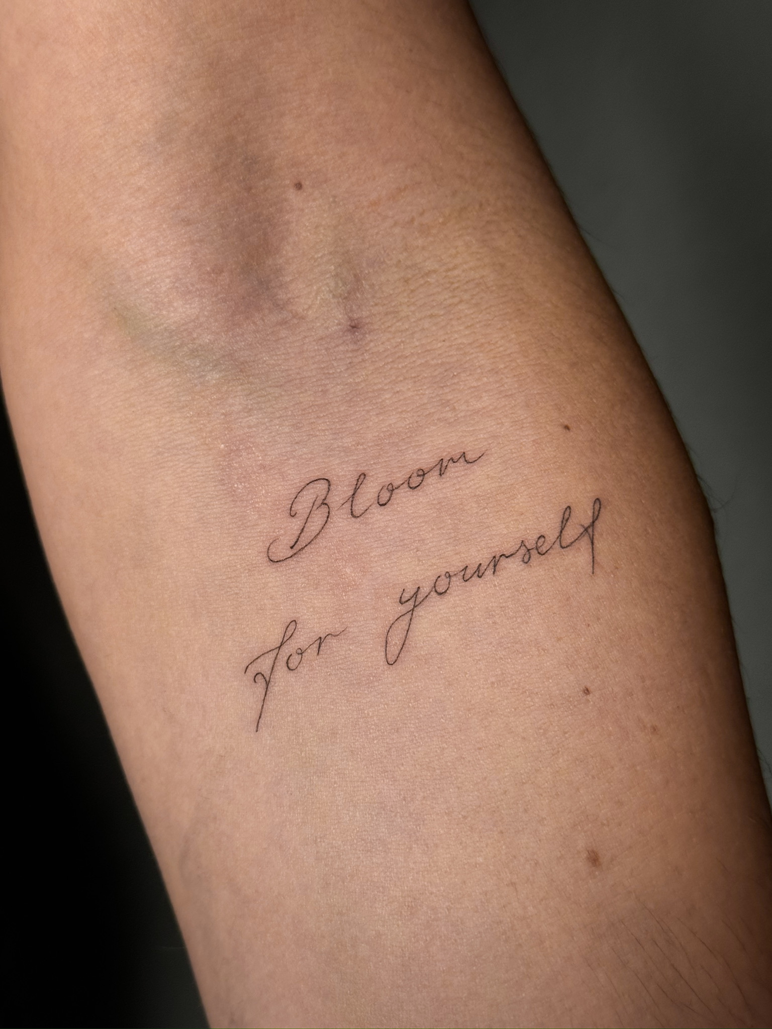 Fine-Line-Tattoo „Bloom for yourself“ in fließender Schreibschrift am Arm.